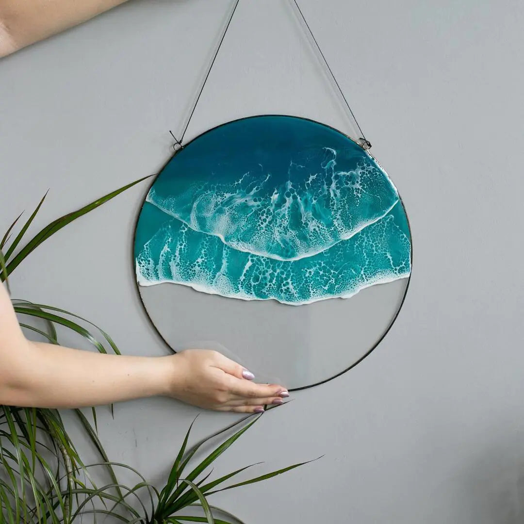 Round ocean wave acrylic wall art with turquoise sea design, hanging on a gray wall and held by a hand with plants around for natural coastal décor vibe. 🌊