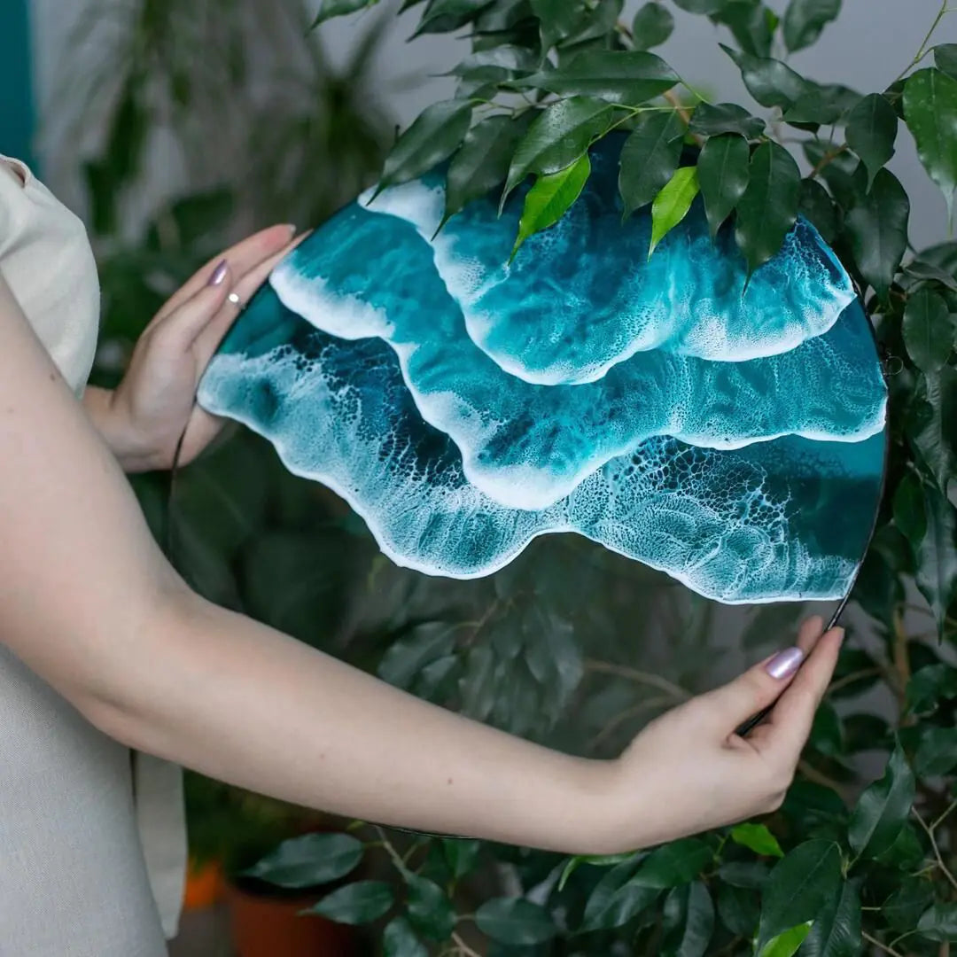 Hand holding ocean wave acrylic wall art among green leaves, showcasing detailed white foam patterns and vibrant blue tones.